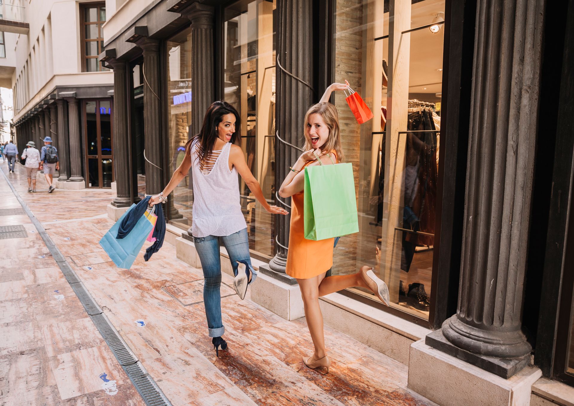 Two women with shopping bags walking excitedly in front of a storefront.