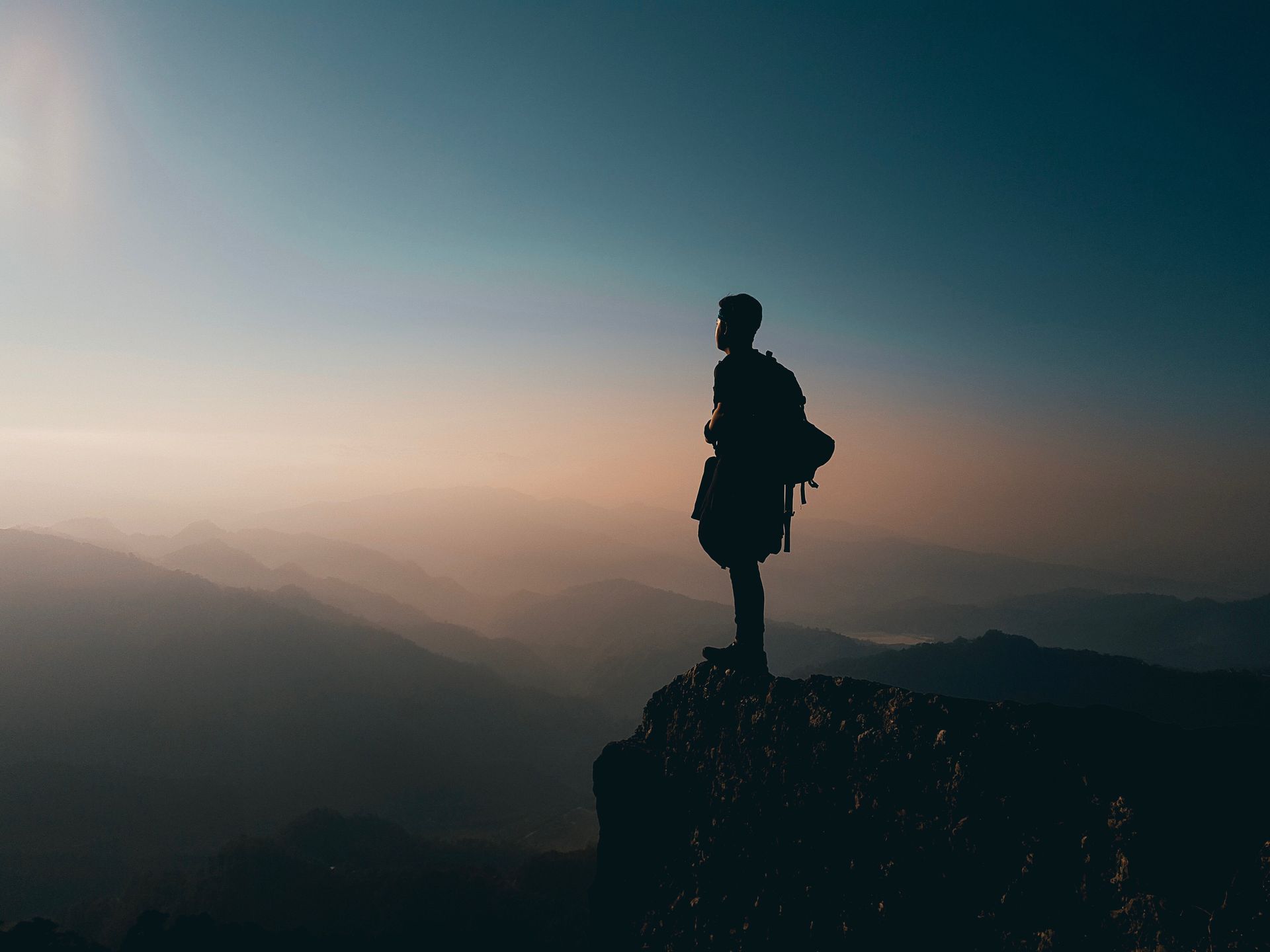 Silhouette of person with backpack on a mountaintop, overlooking layered mountain range at sunrise or sunset.