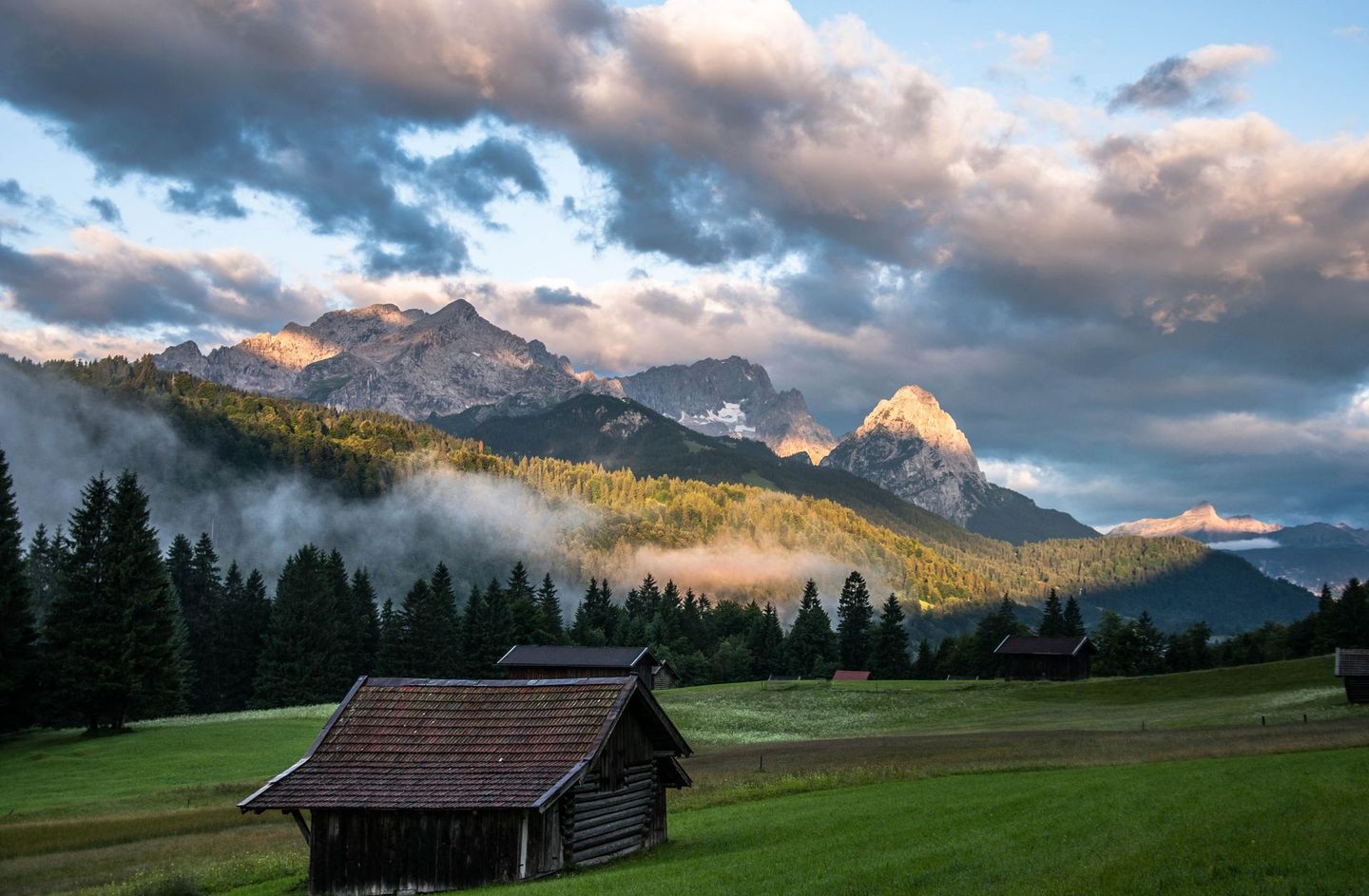 A rustic wooden cabin sits in a lush green meadow before a misty pine forest and dramatic, sunlit mountain peaks.