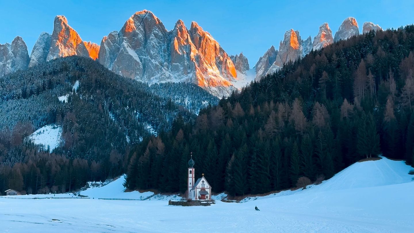 St. Johann Church in the snowy Italian Dolomites at sunset, with golden light hitting the jagged mountain peaks.