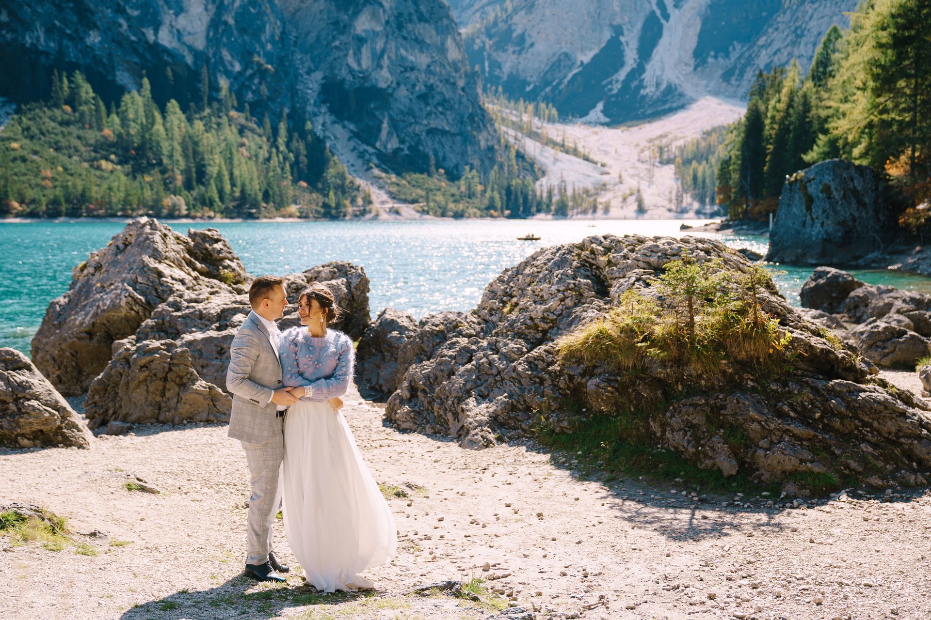 Couple embracing on rocky shore, turquoise lake, mountains in background.