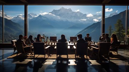 A group sits at a long table, framed by a window overlooking snow-capped mountains and a valley.