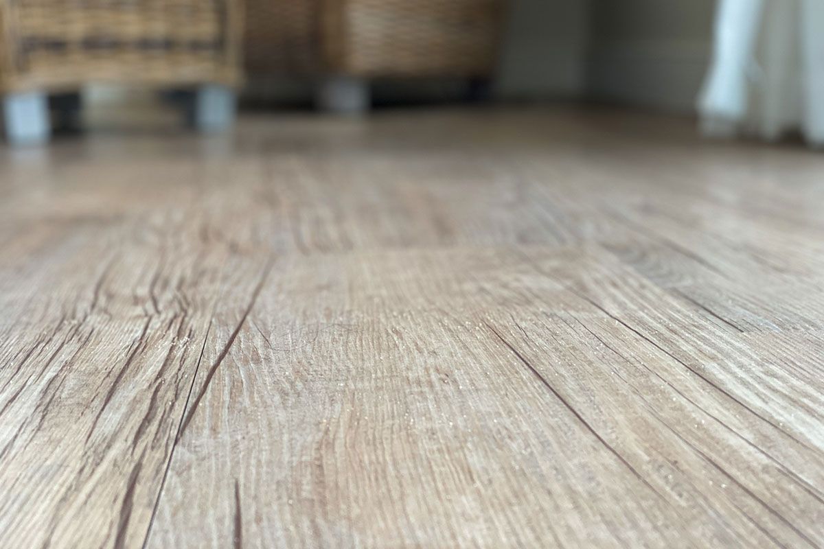 Close-up of light brown wood flooring with subtle grain; out-of-focus furniture in background.