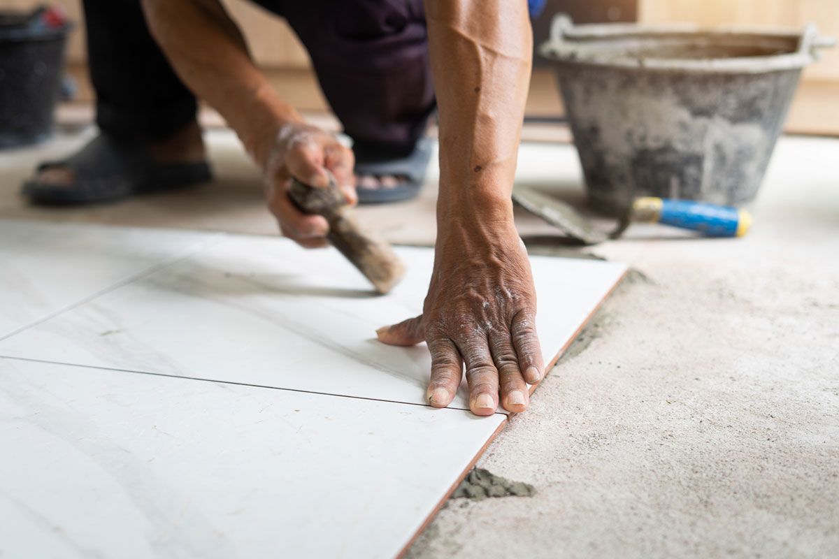 Person tiling a floor, using a trowel. White tile, concrete, bucket in the background.