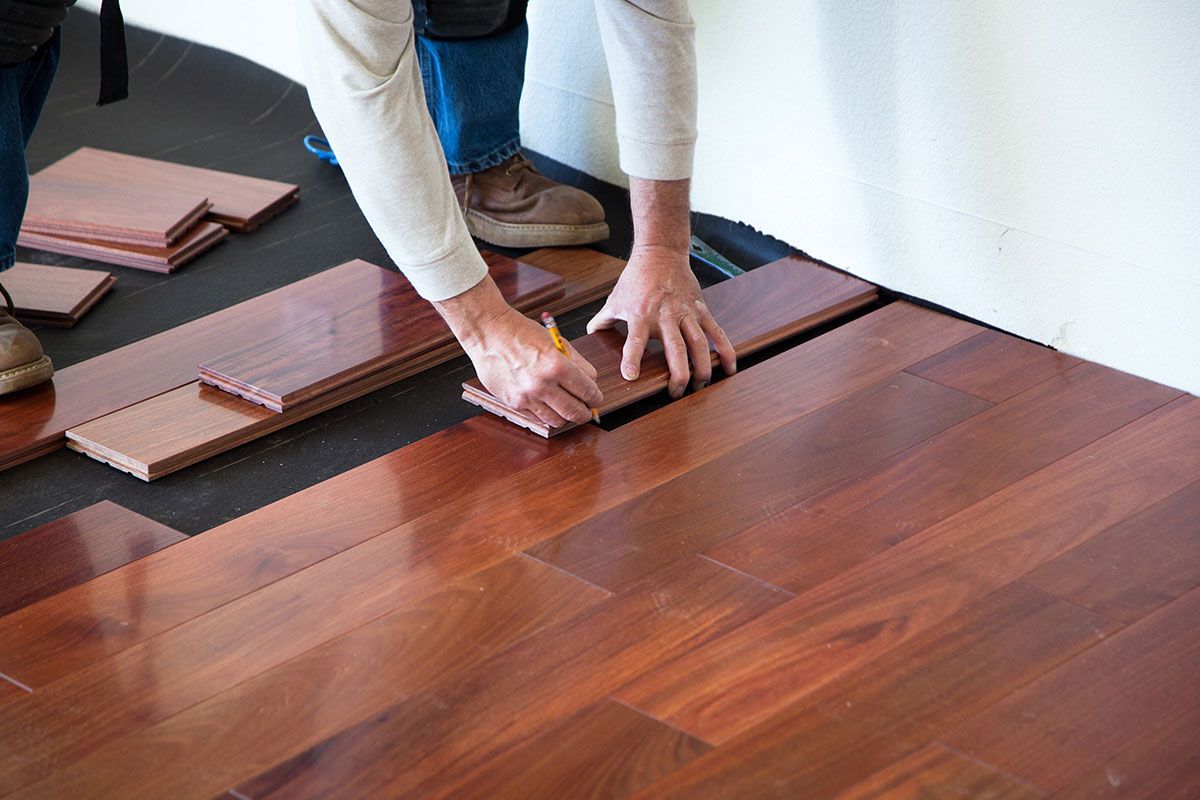 Person installing hardwood flooring; brown wood planks, pencil, measuring.