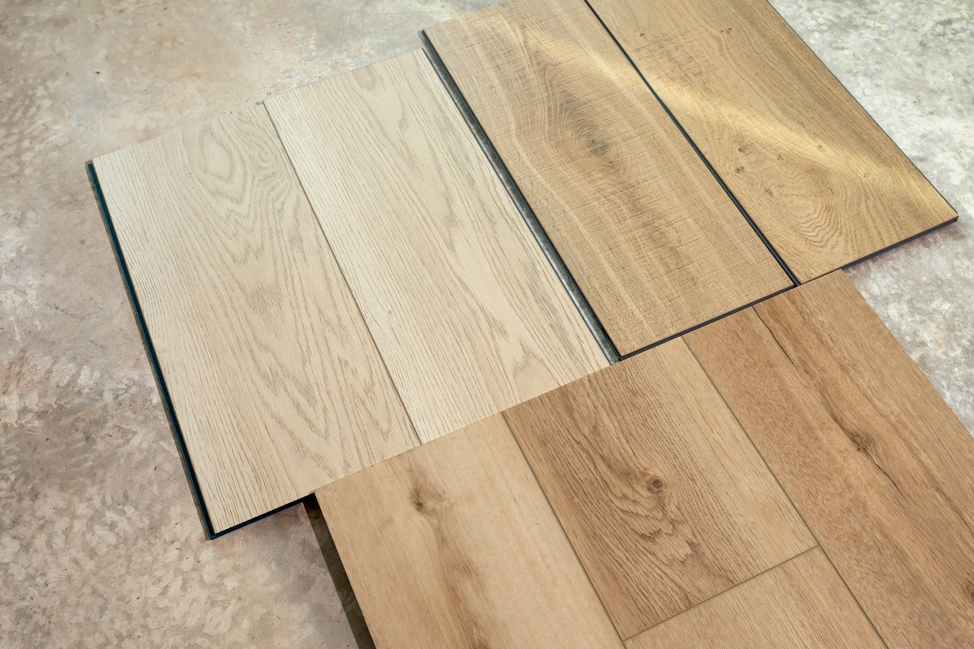 Four wood-look flooring samples in varying light brown and beige tones, arranged on a concrete surface.