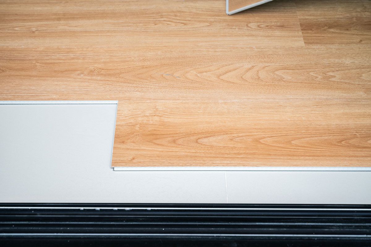 Installing light brown wood-look flooring over white surface, with a black baseboard in the foreground.