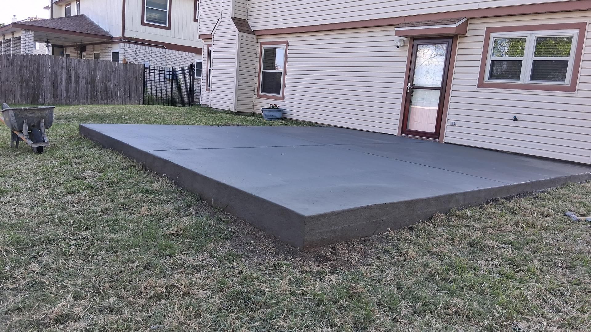 A newly poured, rectangular gray concrete patio sitting in a grassy backyard next to a residential house.