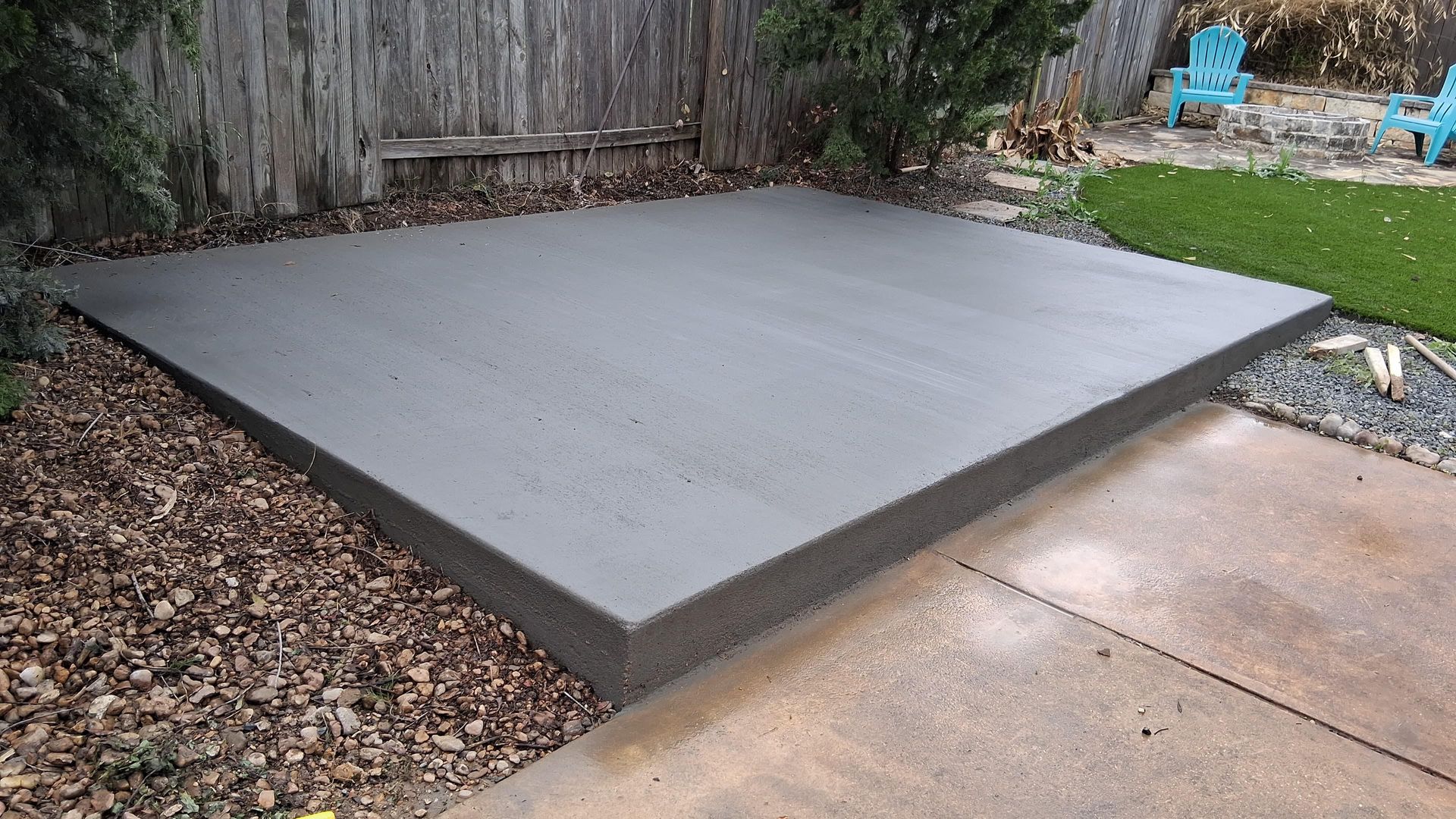 A newly poured, rectangular concrete pad sits in a backyard next to a gravel area and a paved patio.