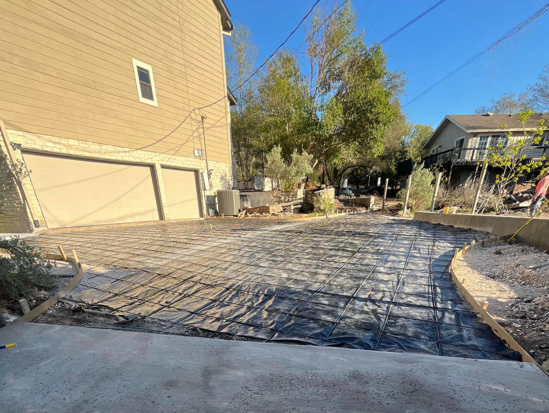 A driveway construction site with a black mesh grid laid over leveled gravel, situated next to a tan house.