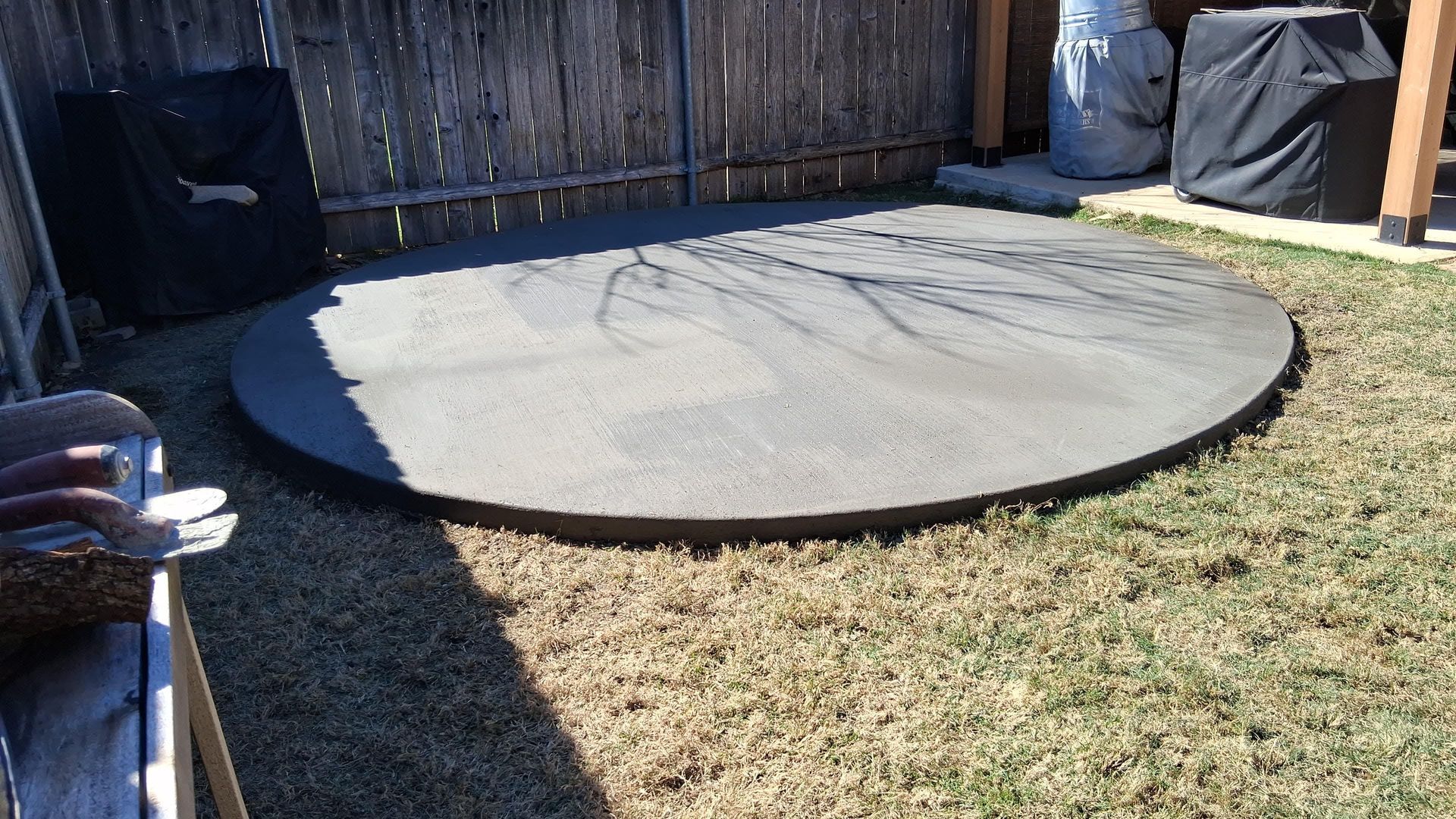 Backyard with brown concrete patio, brown siding, and wooden fence on a sunny day.