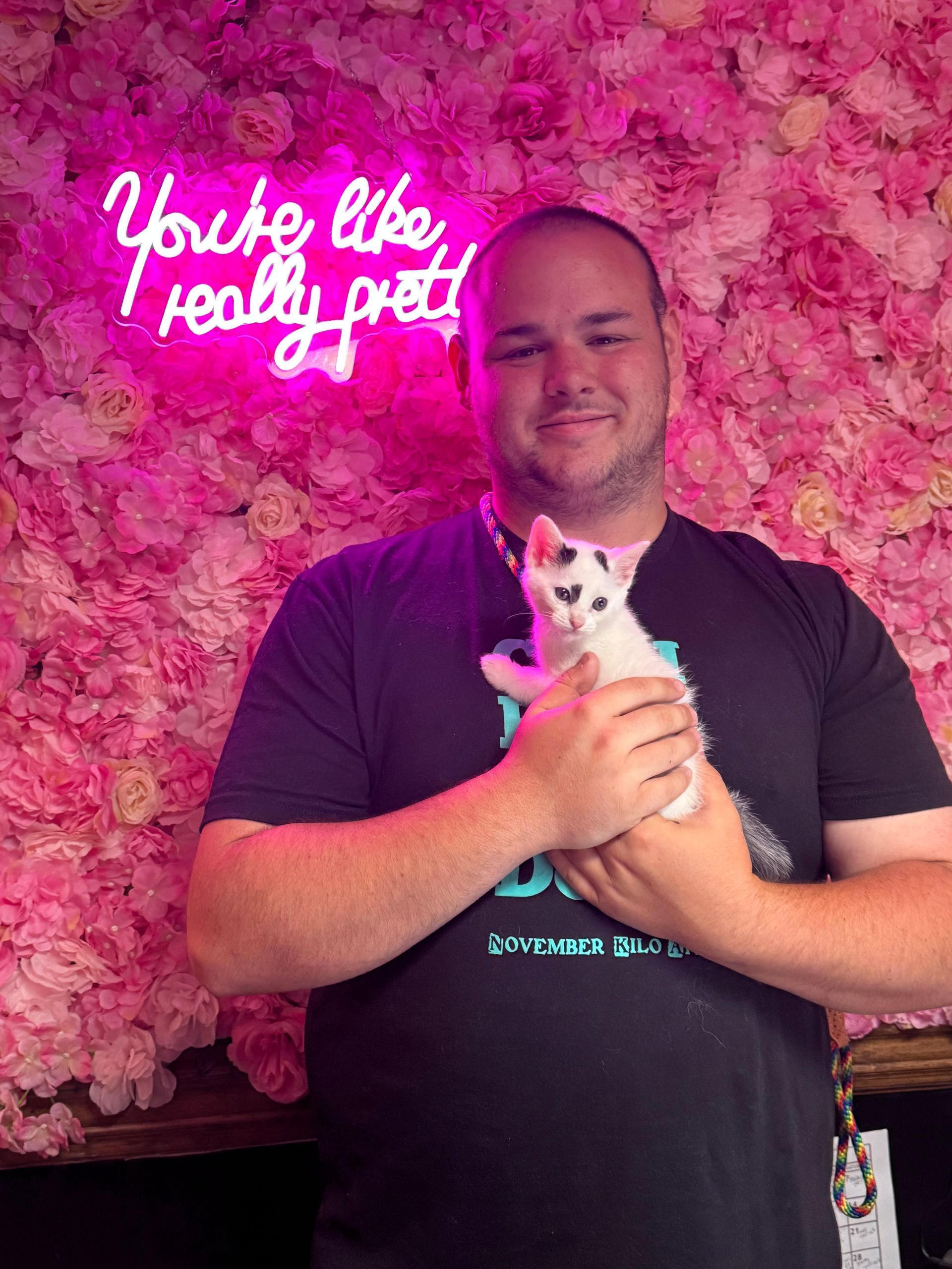A man is holding a kitten in front of a pink flower wall.