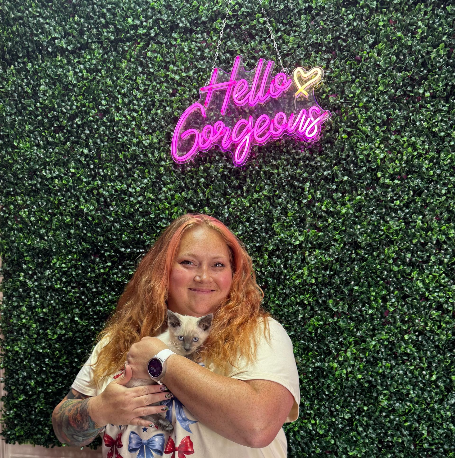 A woman is holding a black cat in front of a wall of pink flowers.