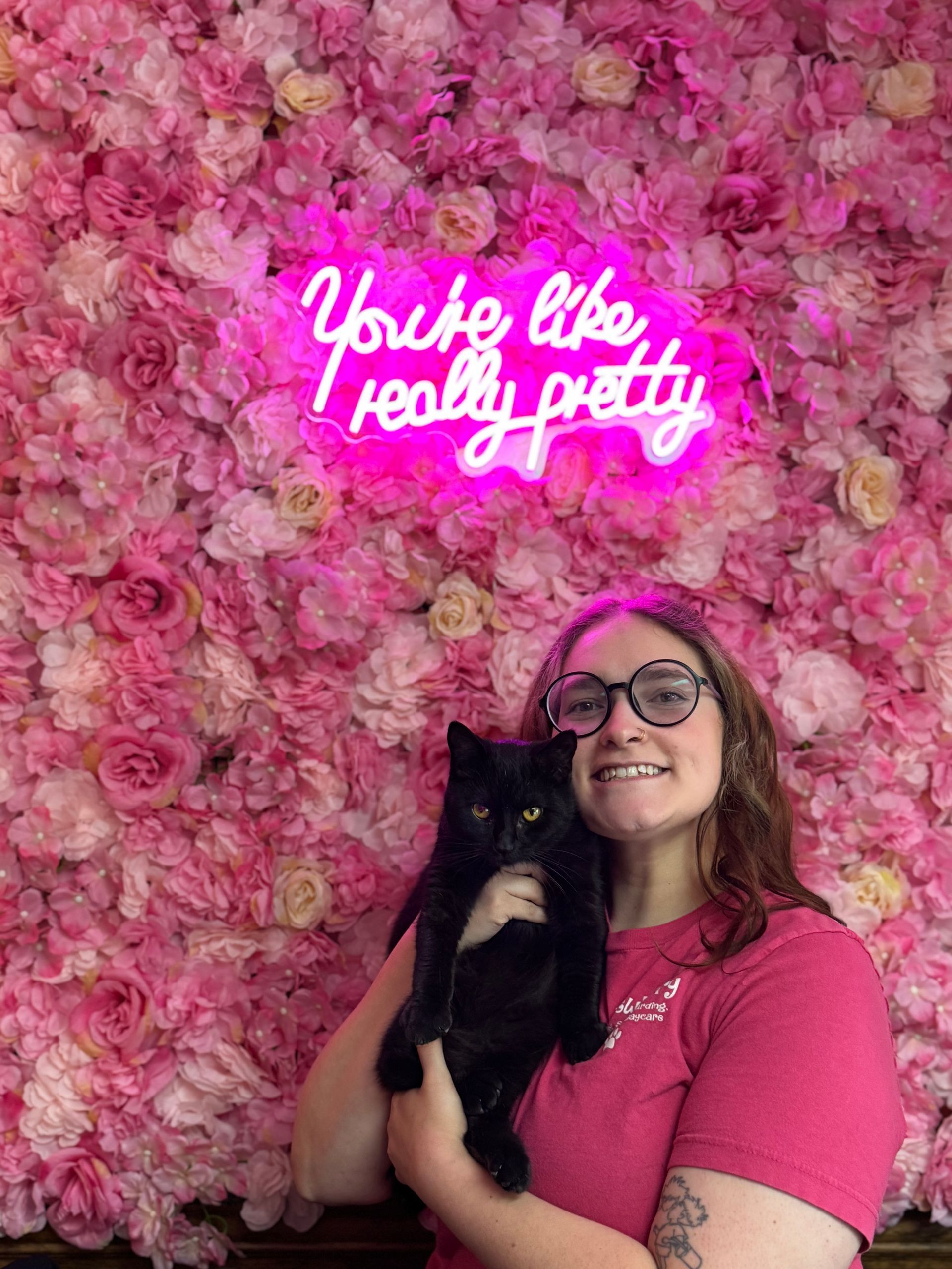 A woman is holding a black cat in front of a wall of pink flowers.