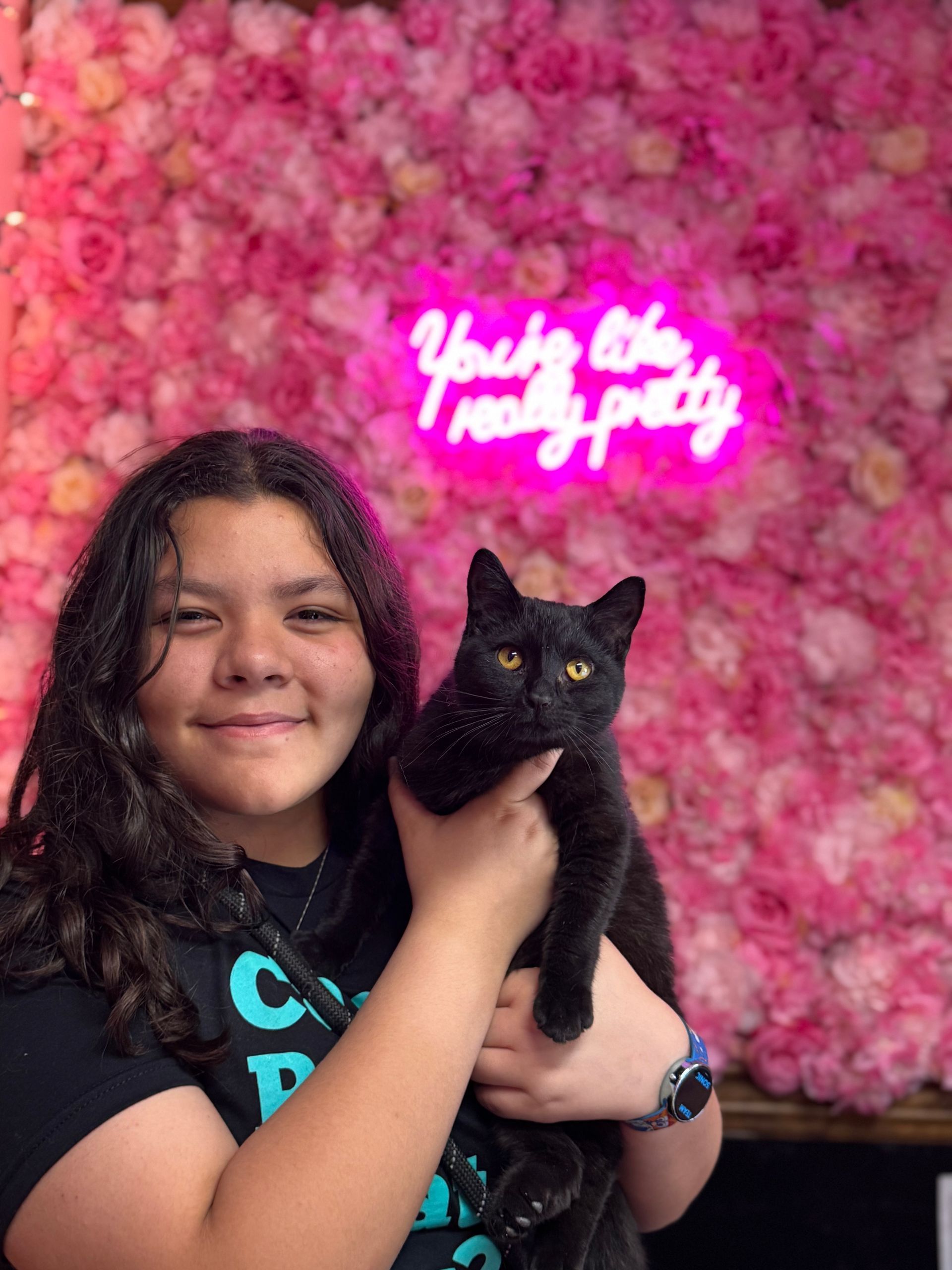 A girl is holding a black cat in front of a pink flower wall.