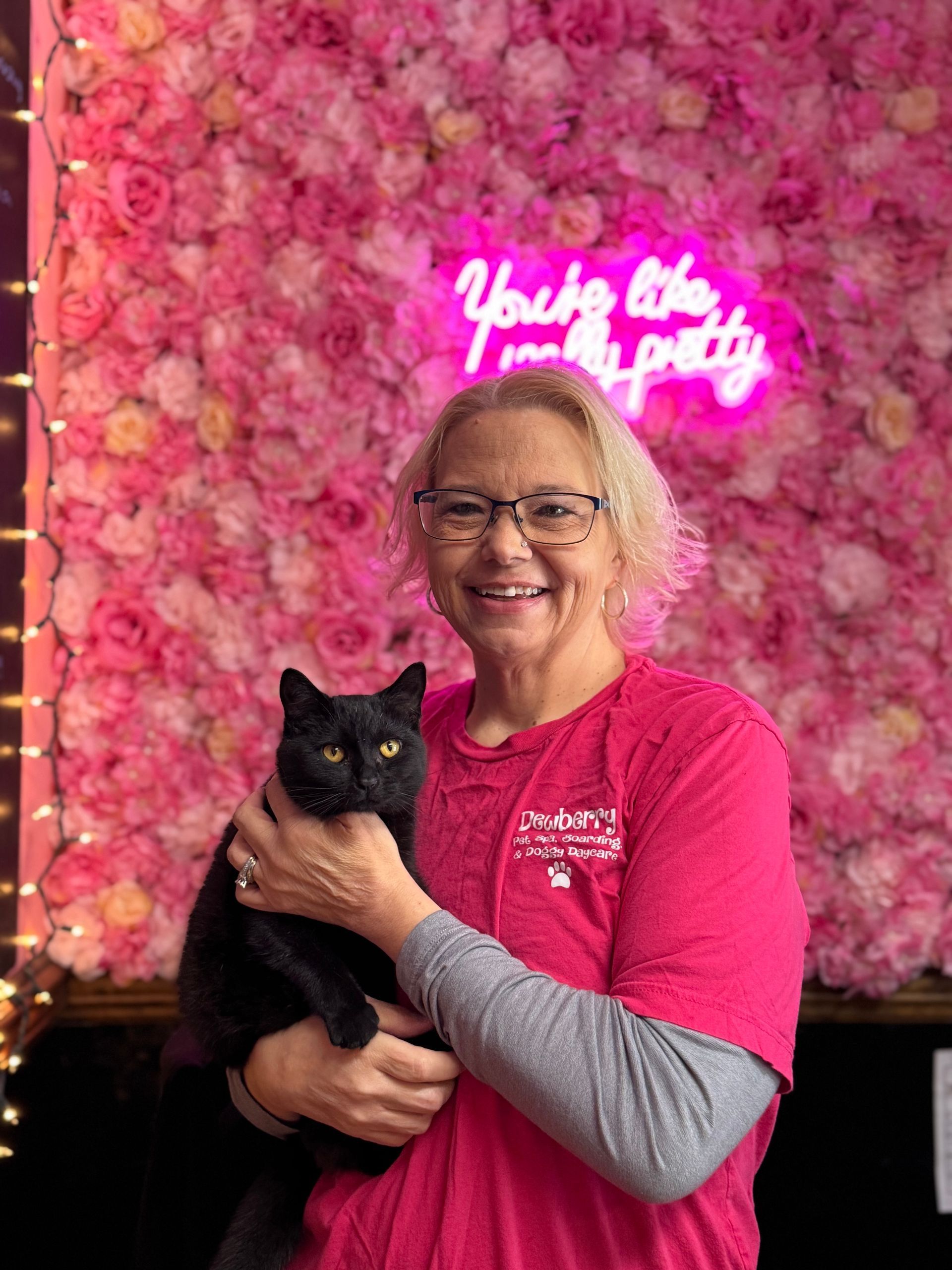 A woman in a pink shirt is holding a black cat in front of a wall of flowers.