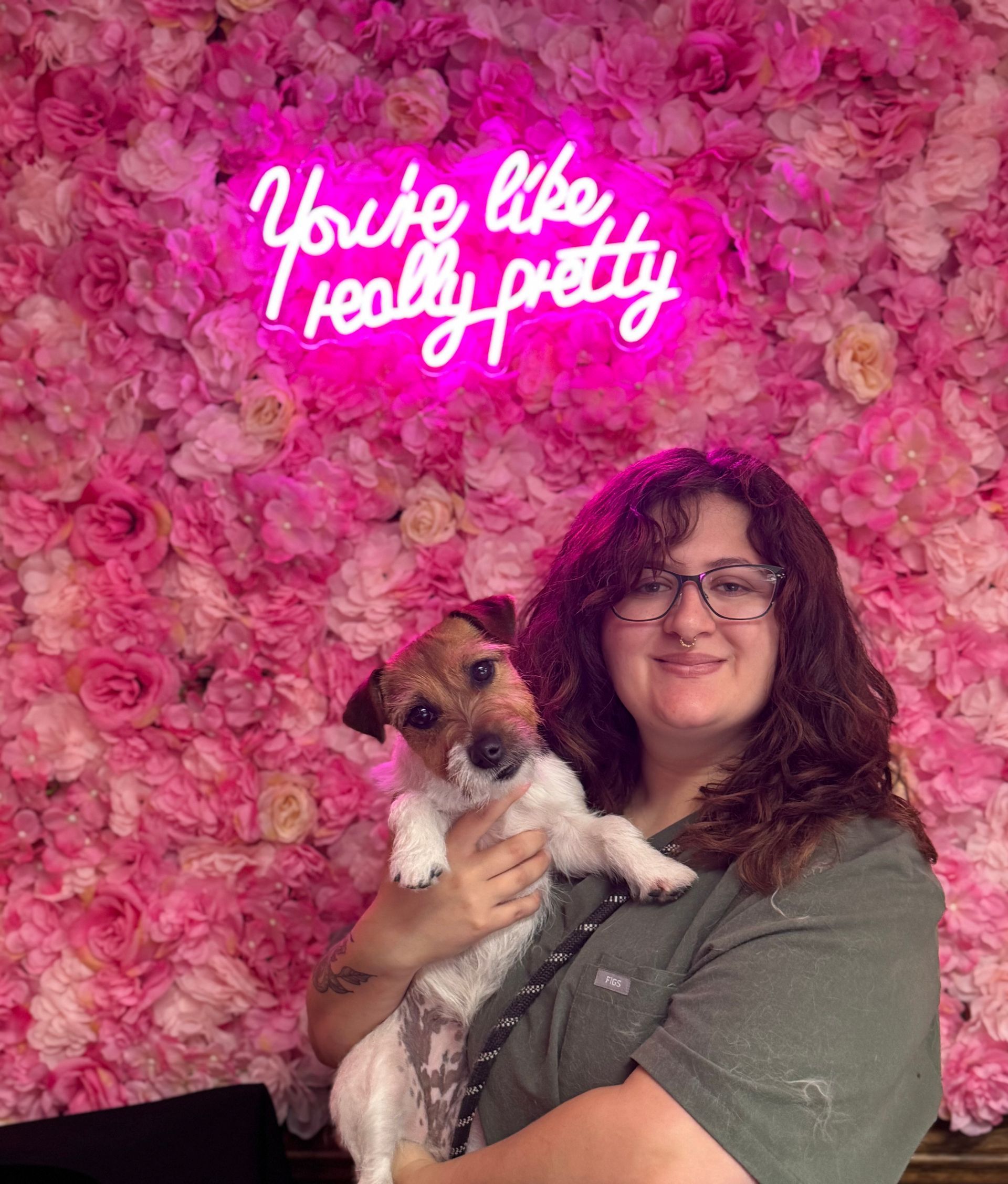 A woman is holding a small dog in front of a pink flower wall.