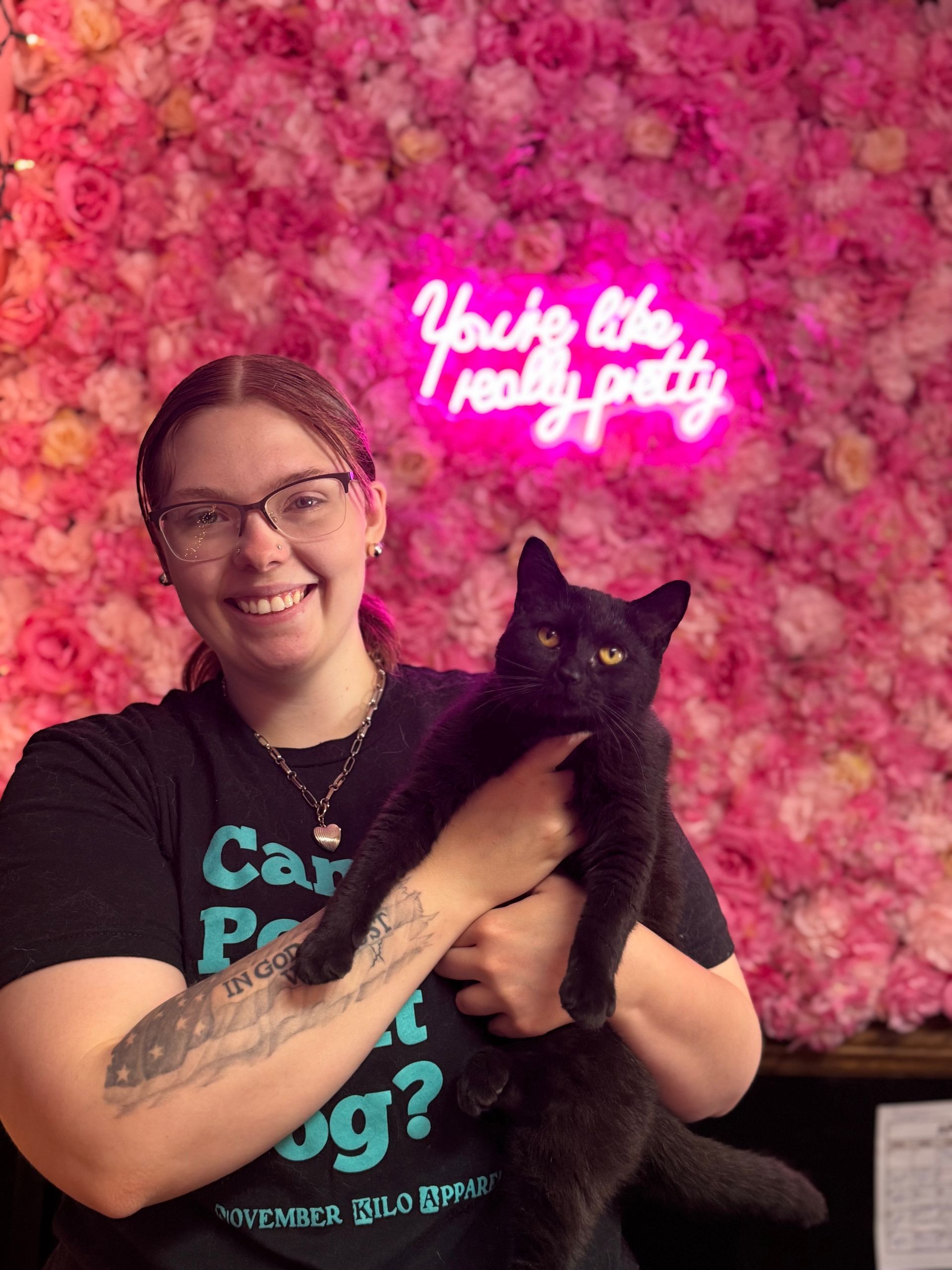 A woman is holding a black cat in front of a pink flower wall.