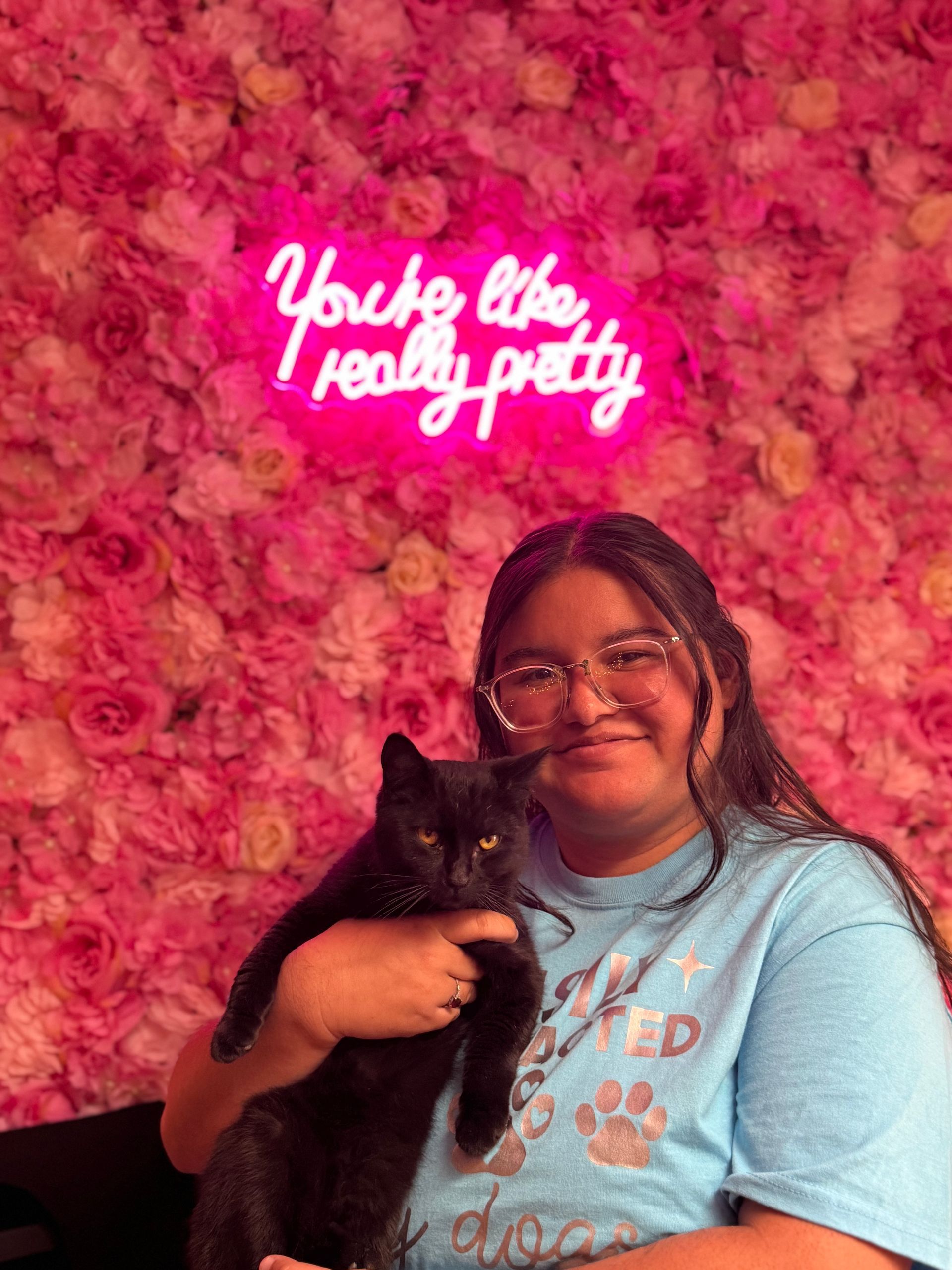 A woman is holding a black cat in front of a pink flower wall.