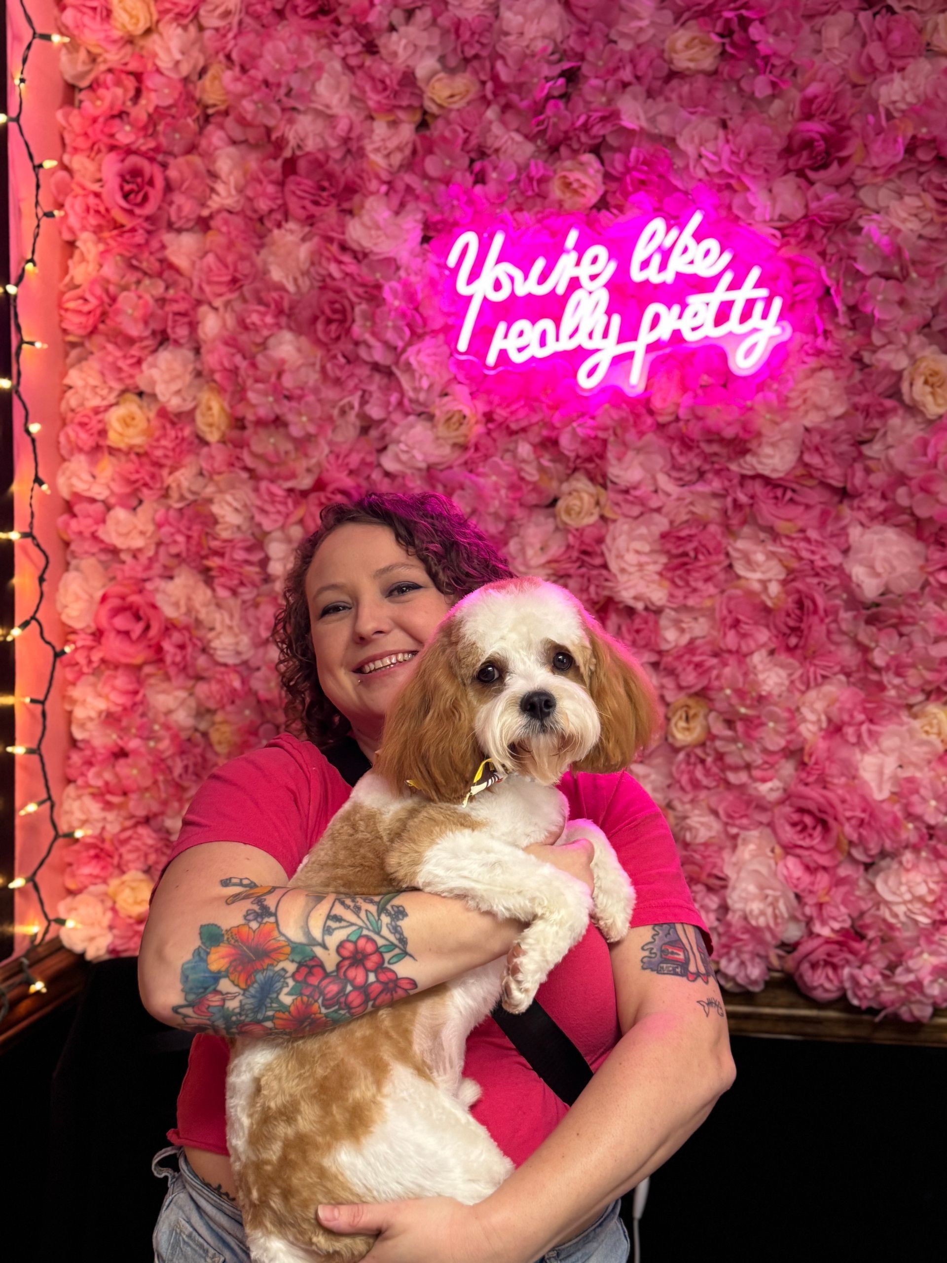 A woman is holding a small dog in front of a pink flower wall.