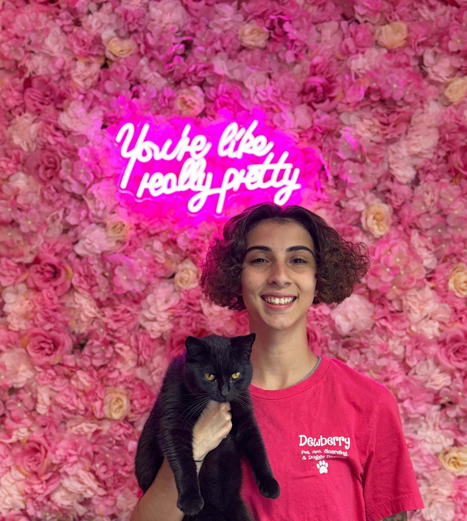 A young man is holding a black cat in front of a wall of pink flowers.
