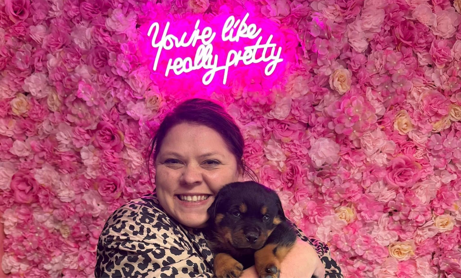 A woman is holding a puppy in front of a pink flower wall.