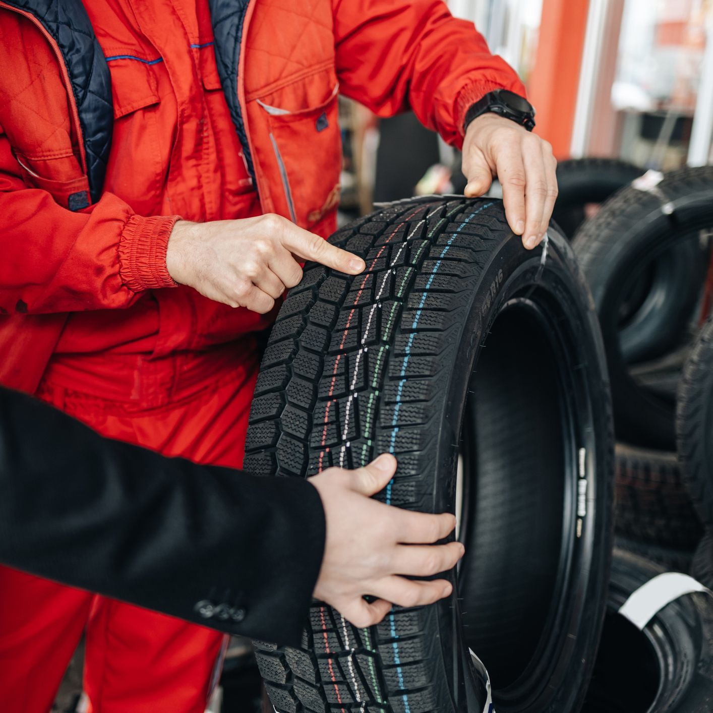 Mechanic pointing at tire tread; a customer in a black suit holds the tire in a shop.