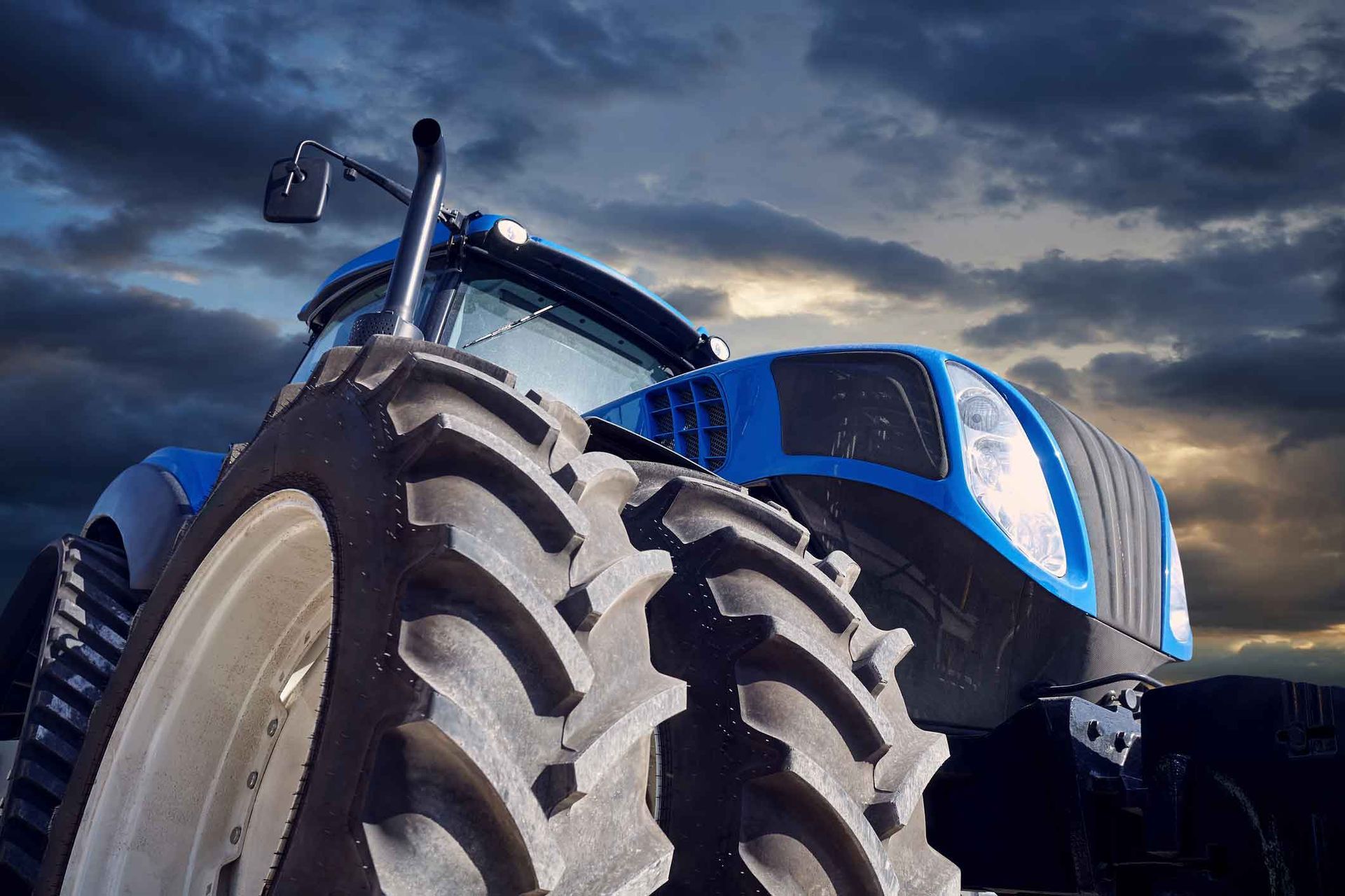 A blue tractor is parked in a field with a cloudy sky in the background.
