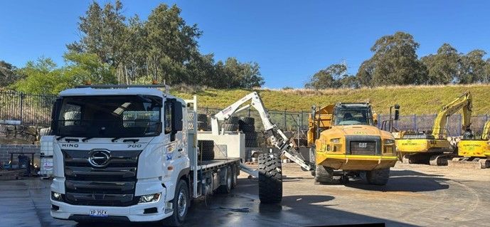 A white truck with a crane loading a large black tire next to yellow construction equipment. Sunny day.