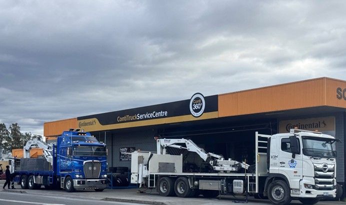 Blue semi-truck and white service truck parked outside an orange and black building, under a cloudy sky.