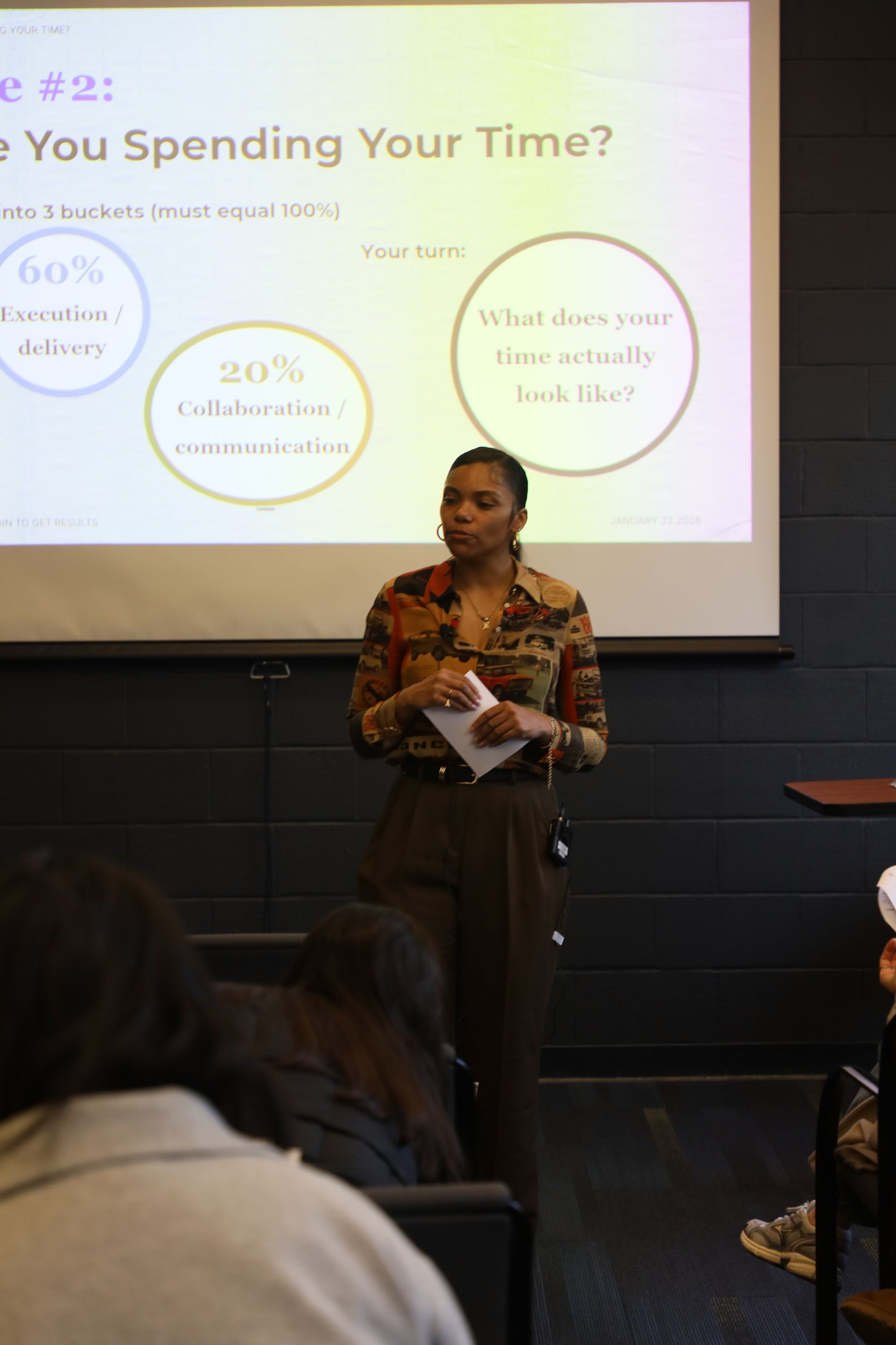 Man giving a presentation to a group; he’s holding a tablet, and the projector displays a slide with text.