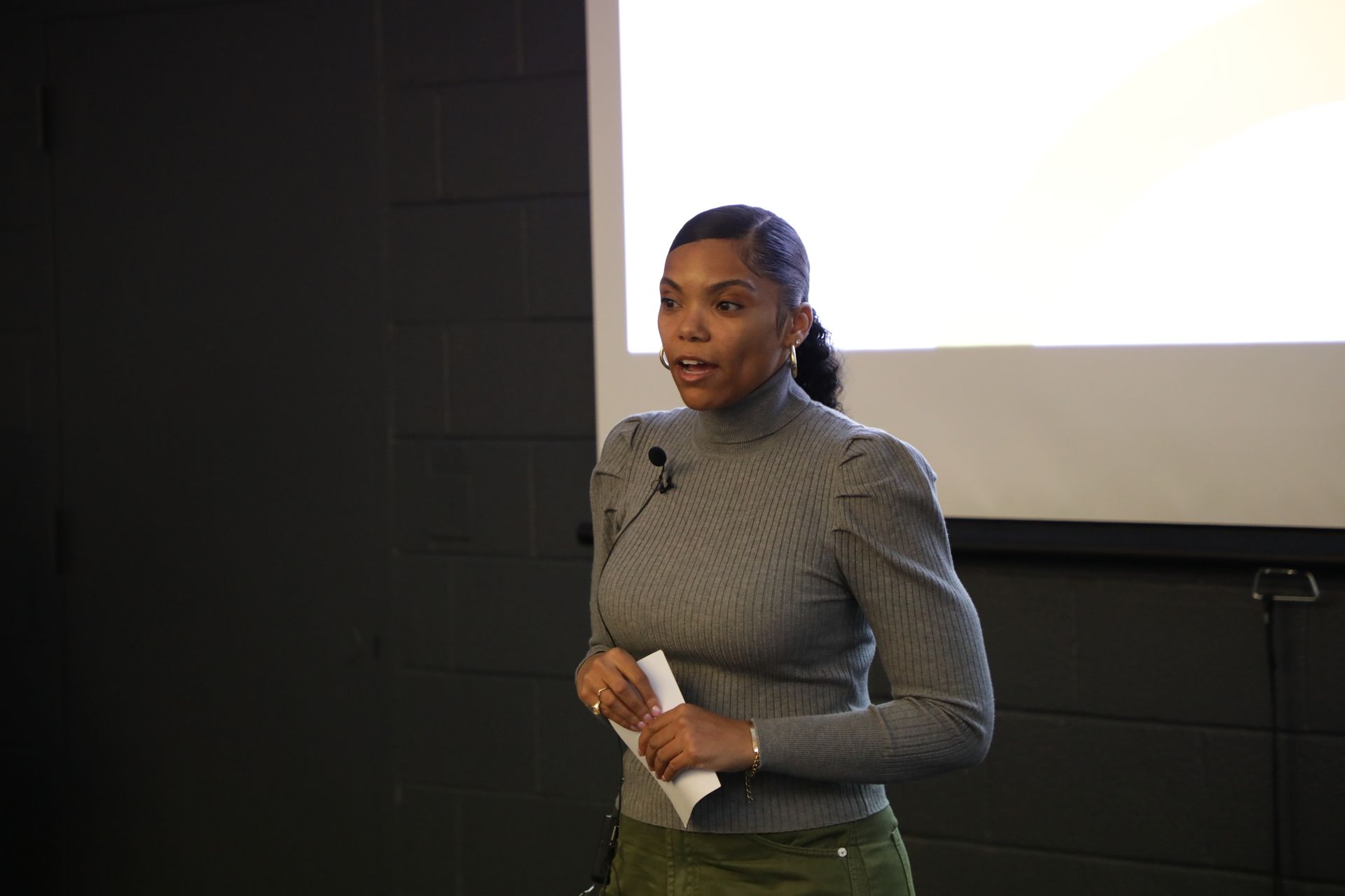 Four people in a meeting, one presenting, others looking at documents. A projector screen is behind.