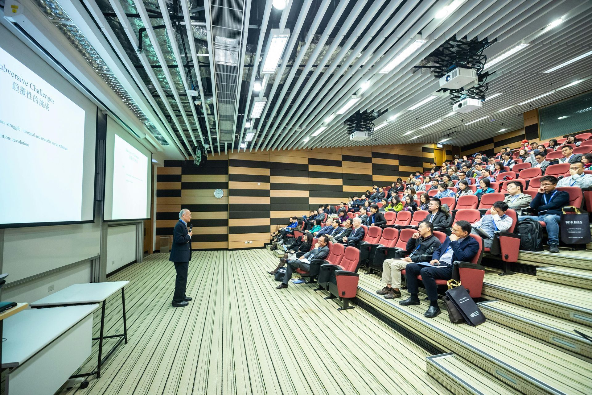 Professor lecturing to a large class in a tiered auditorium. Projection screens show text.