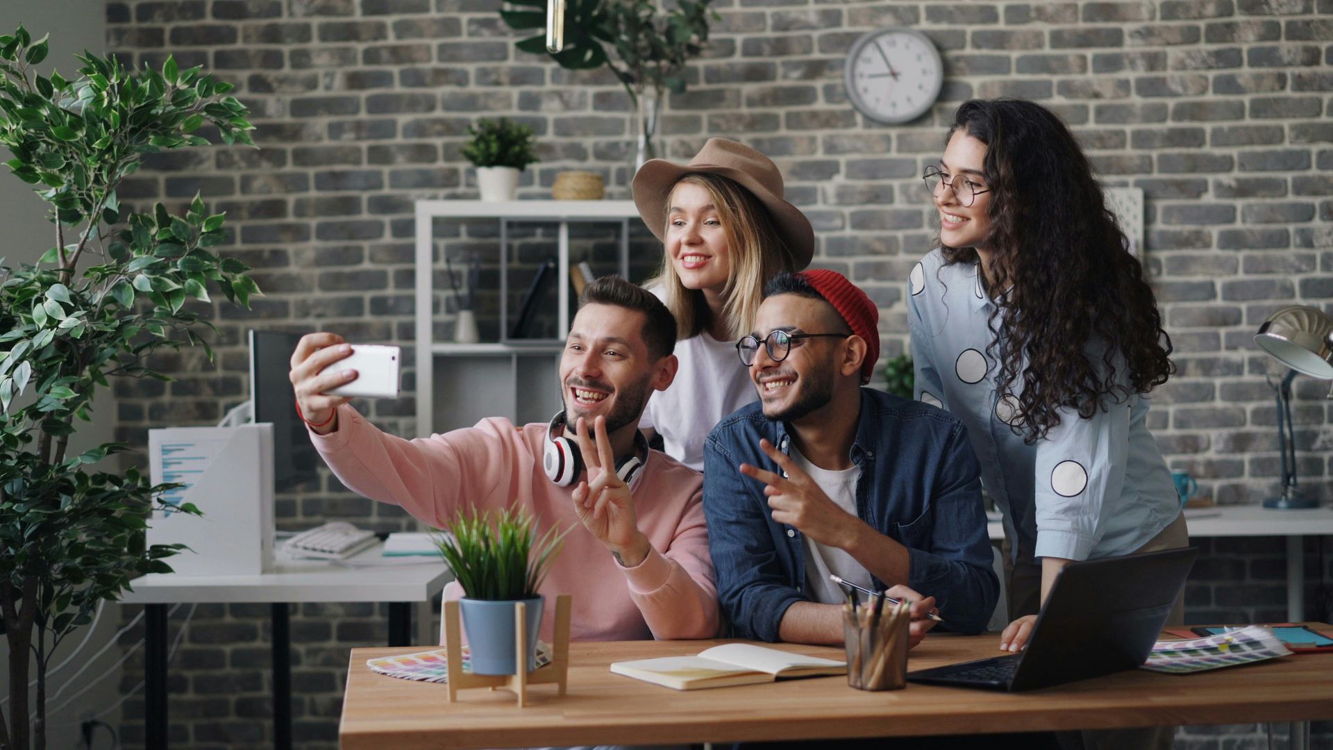 Four people taking a selfie in an office; two men and two women smiling, posing, and using a smartphone.