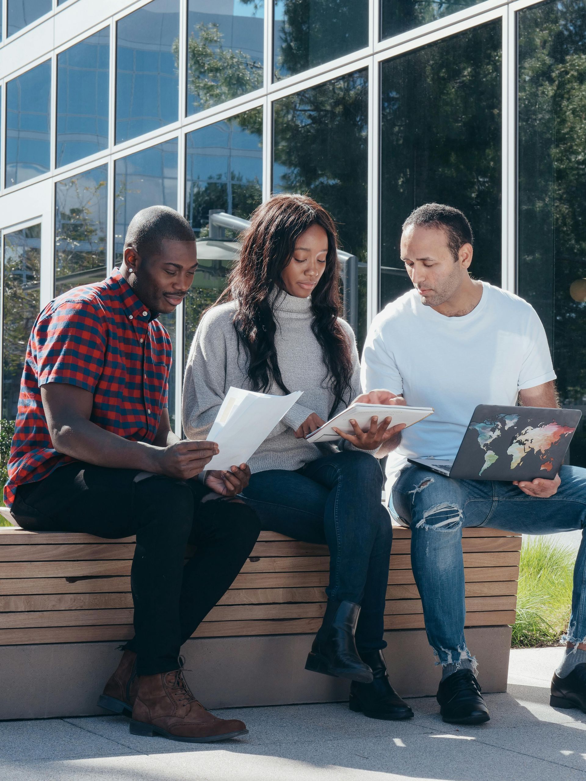 Three people seated on a bench, looking at papers and a laptop in front of a modern building.