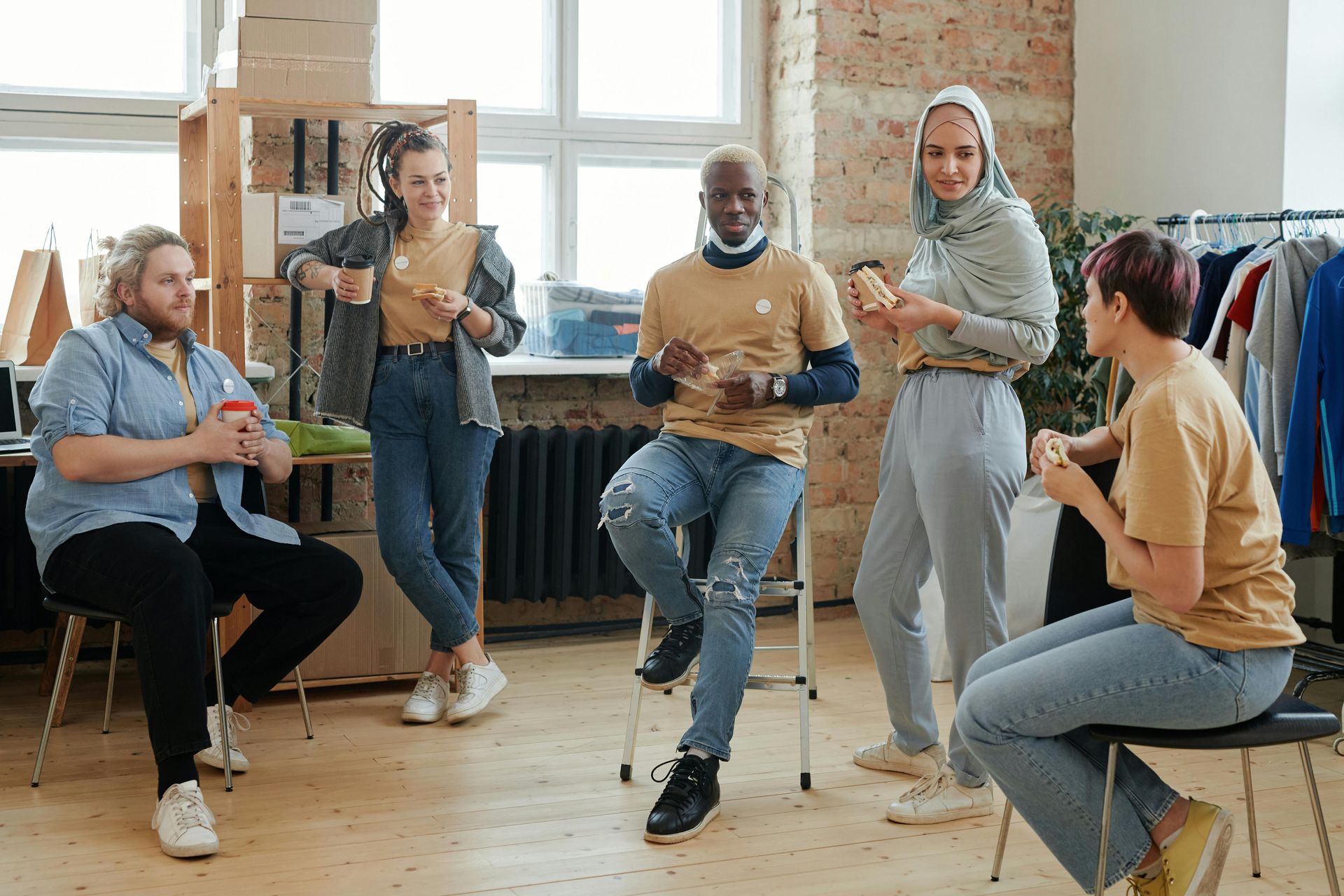 Group of people in casual attire, conversing while eating. They're indoors, relaxed, and near clothing racks.