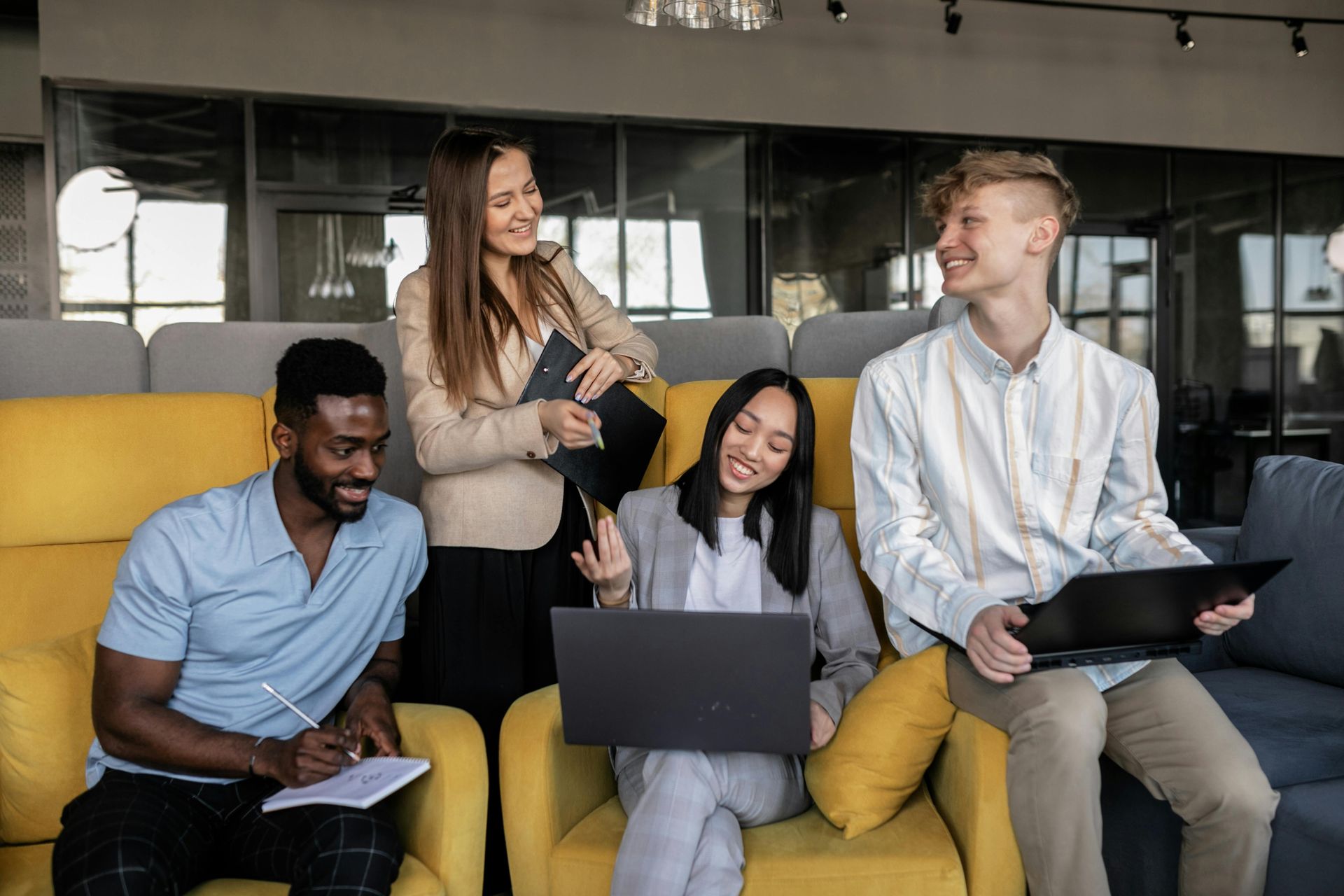 Four colleagues in an office setting; two on yellow armchairs, two standing. All are interacting, smiling, and using laptops or notebooks.