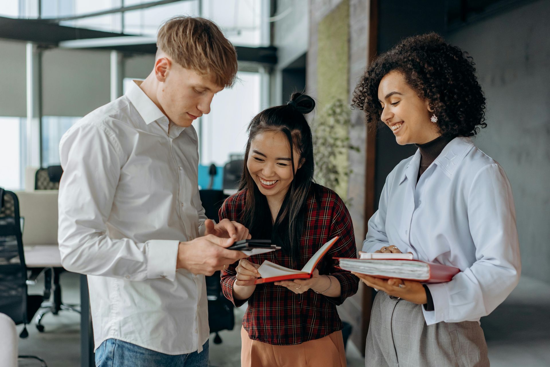Three people in office, looking at phone and notebooks, smiling.