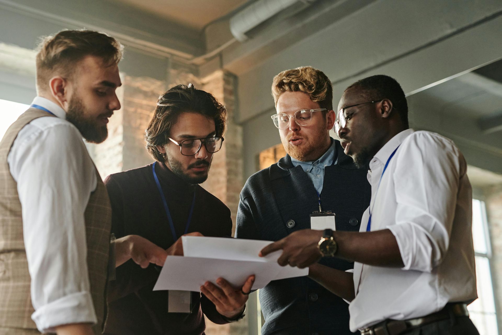 Four colleagues in a well-lit room reviewing documents together. They are wearing lanyards and engaged in conversation.