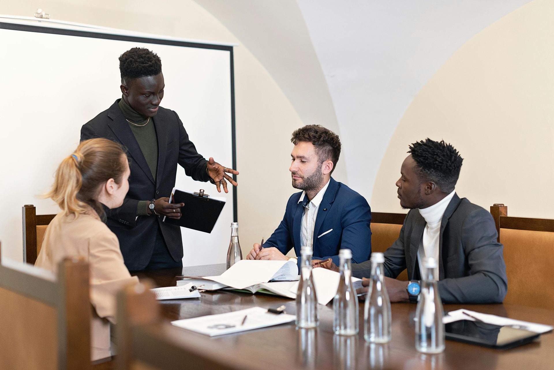 Four people in a meeting, one presenting, others looking at documents. A projector screen is behind.