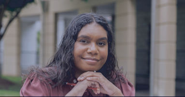 A young woman is sitting in front of a building with her hands folded and smiling.