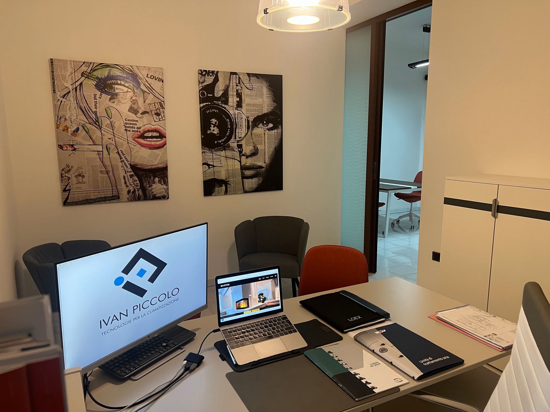 Scrivania da ufficio con computer portatile, monitor con logo, opera d'arte sulla parete e porta aperta.