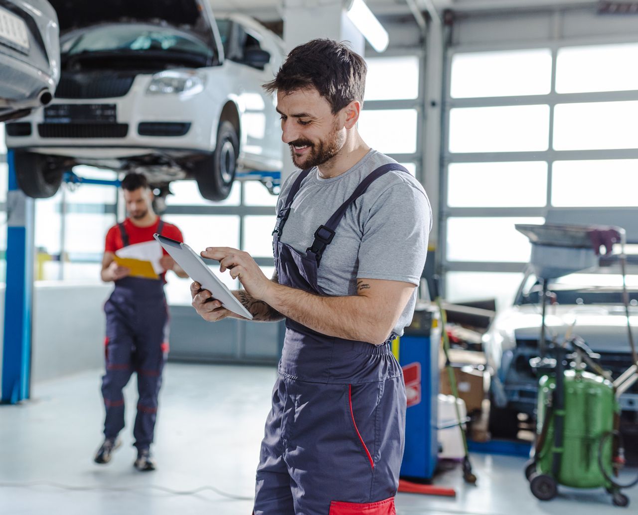 A man is using a tablet in a garage while another man looks at a clipboard.