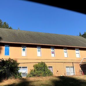 Two-story building with tan siding, several windows, and a dark roof under a blue sky.