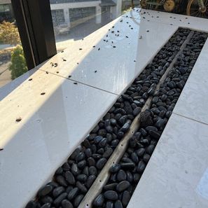 A long, rectangular fire pit with black rocks, set in white tile. Black debris is scattered on the tile.