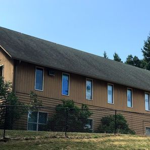 Tan building with multiple rectangular windows, dark roof, behind a black fence.