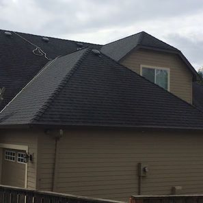 Dark shingled roof on a two-story tan house, with a dormer and garage. Overcast sky.
