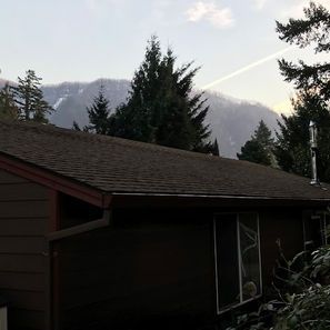 Brown house with dark roof and mountain backdrop. Trees in front. White contrail in sky.