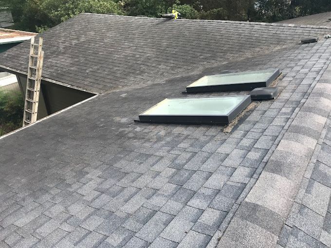 Gray asphalt shingle roof with two skylights and a ladder on the side.