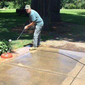 Man using a circular surface cleaner to power wash a concrete patio outdoors on a sunny day.